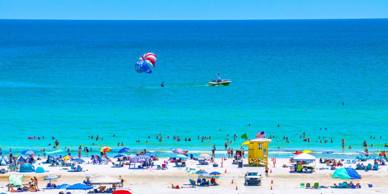 Many beachgoers relax on Siesta Key while paragliders soar above them against a sunny backdrop.