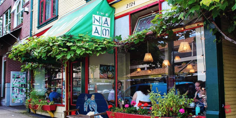 The Naam restaurant in Vancouver. It has a green awning, large windows, and outdoor planters with flowers and greenery. Inside, people are seated at tables.