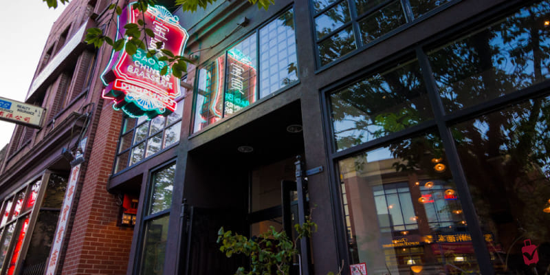 The exterior of "Bao Bei Chinese Brasserie" restaurant. It features a bright neon sign with Chinese characters and the restaurant name, large windows, and brick architecture.