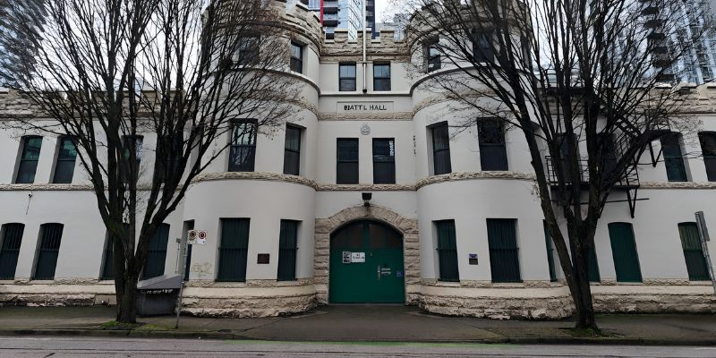 Facade of the Beatty Street Drill Hall, a historic white building resembling a castle with crenelated turrets, a large arched green door, and leafless trees in the foreground.