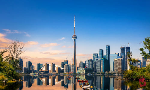 The Toronto skyline is reflected in the calm waters of a lake, with the CN Tower prominently featured among numerous skyscrapers, and small boats with people are visible on the water, all under a clear blue sky with a hint of sunset colors on the horizon.