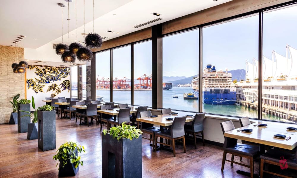 A modern restaurant dining room with large windows overlooking a harbor with ships, cranes, and waterfront buildings. Tables are set, and decorative elements include plants and unique light fixtures.