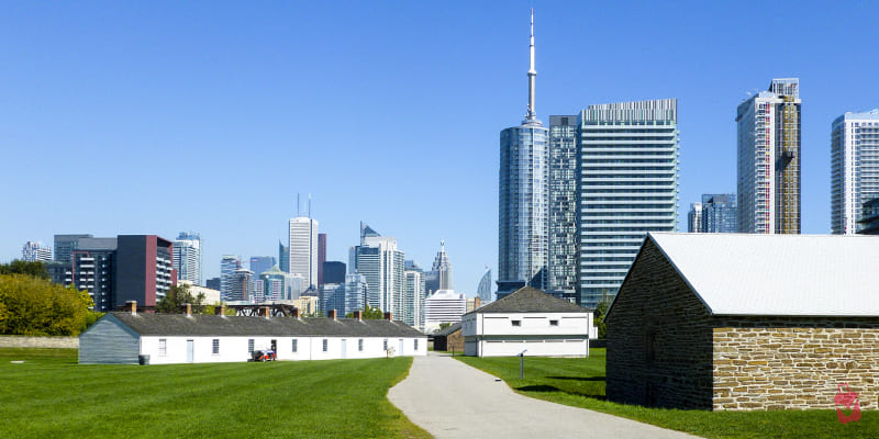 Buildings of Fort York, a historical site with a grassy path leading towards them, set against the backdrop of the modern Toronto skyline under a bright blue sky.