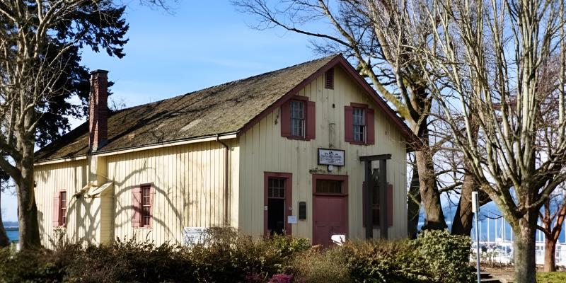 The Old Hastings Mill Store Museum, a small, historic pale-yellow wooden building with a shingled roof and a red brick chimney, surrounded by bare trees and grass.