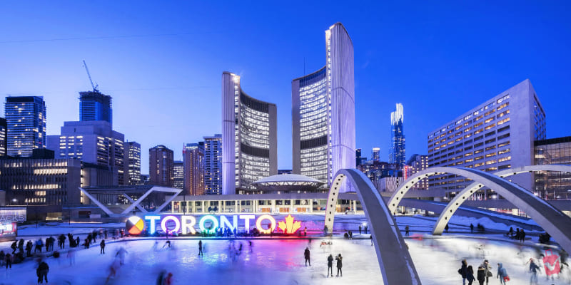 The illuminated "TORONTO" sign in Nathan Phillips Square at dusk, with people ice skating and modern city buildings lit up in the background.