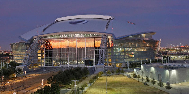 An exterior view of AT&T Stadium at dusk, with the building's lights reflecting on its facade.