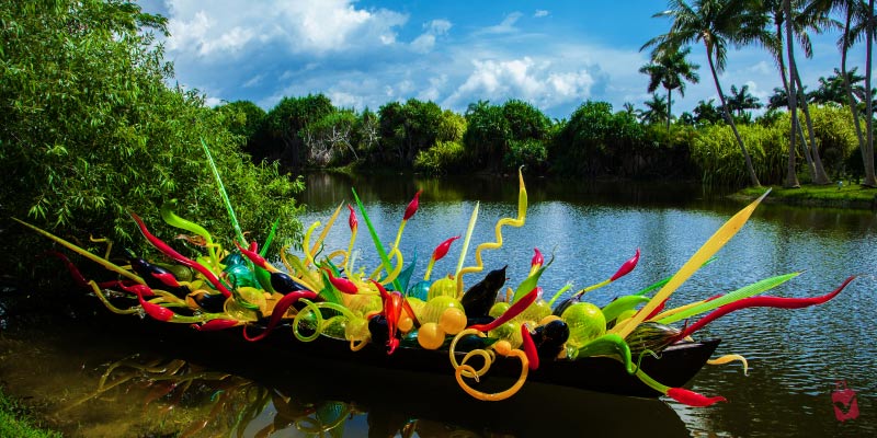 A colorful glass sculpture in the shape of a boat floating on a body of water, surrounded by lush greenery.