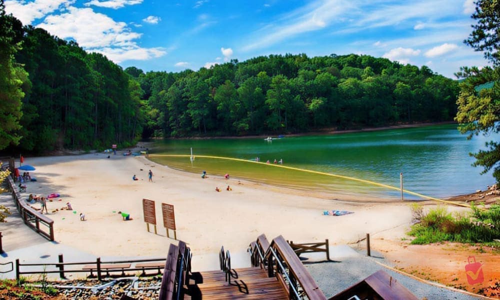 A gorgeous sunny day at this Atlanta beach! Folks are happily enjoying the sand and swimming in the calm lake, beautifully framed by lush green trees.
