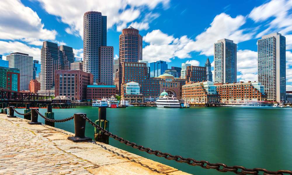 Waterfront view of the Boston skyline with modern skyscrapers, a cobblestone harbor walk, and clear blue skies with wispy clouds.