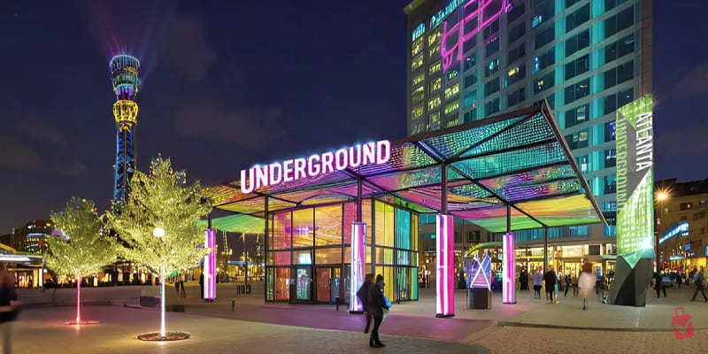 Vibrant night view of the Underground Atlanta entrance with colorful neon lights, a modern building, and the lighted Atlanta Tower in the background.