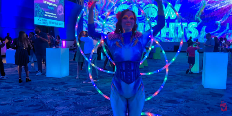 Performer in a vibrant blue costume with neon body paint, holding LED hoops, at a Georgia Aquarium event. Blue lighting fills the lively party scene.