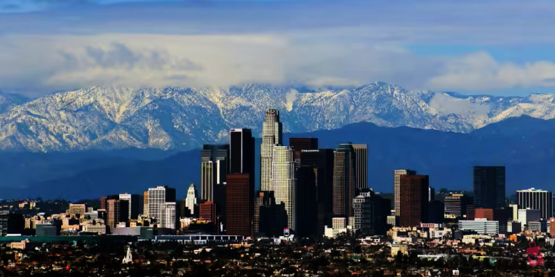 The Los Angeles skyline with tall buildings against a backdrop of snow-capped mountains and a cloudy sky.