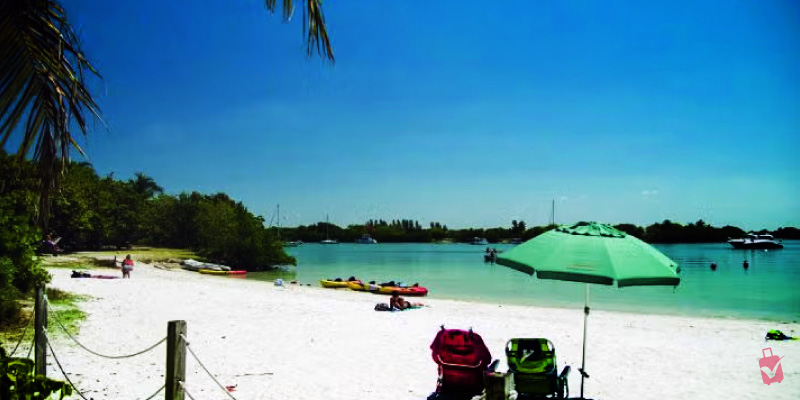 A sunny beach scene at Oleta River State Park with people relaxing, kayaks on the sand, and boats in the water.