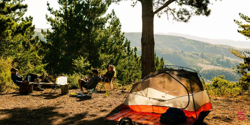 People relaxing around a tent and campfire in a scenic, forested area, showcasing the Portolá Trail Campsites experience.