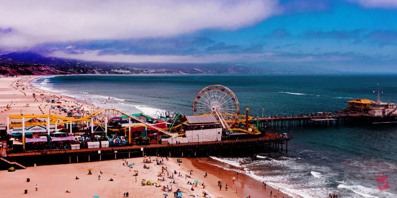 An aerial view of Santa Monica Pier and beach, with a Ferris wheel and roller coaster under a cloudy sky.