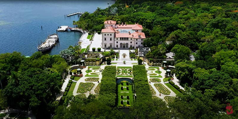 An aerial view of the Vizcaya Museum and Gardens, showcasing a large mansion and elaborate formal gardens beside the water.