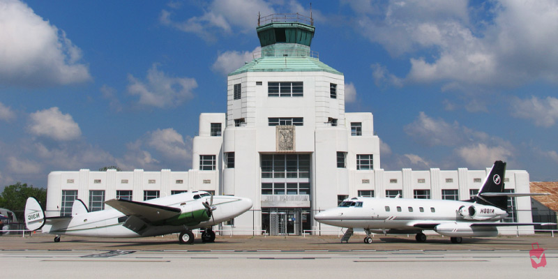 Two vintage aircraft sit parked in front of the historic white art deco building of the 1940 Air Terminal Museum under a bright blue sky.
