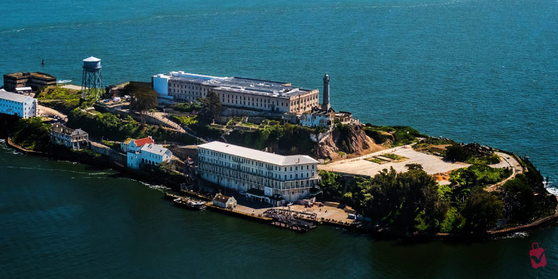 An aerial view of Alcatraz Island showing the historic prison buildings and lighthouse surrounded by the deep blue waters of the San Francisco Bay.