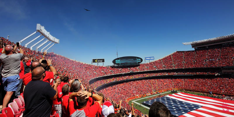 Fans in red fill Arrowhead Stadium as a stealth bomber flies over a massive American flag on the field during an exciting game day celebration.