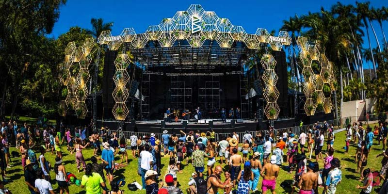 An outdoor music festival with a band performing on a stage and a crowd of people dancing on a grassy field surrounded by palm trees.