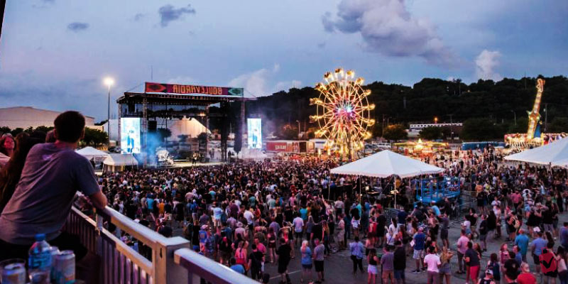 A massive crowd gathers at Boulevardia for an outdoor music festival featuring a large illuminated stage and a glowing Ferris wheel at dusk.
