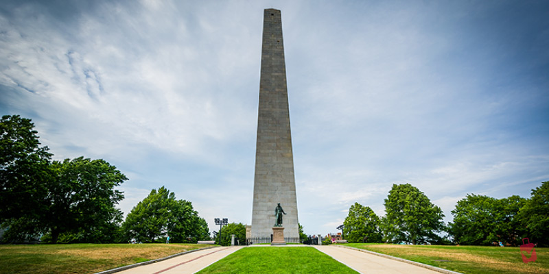 The tall granite Bunker Hill Monument obelisk stands prominently against a cloudy sky, marking a significant site of American Revolutionary history in Boston.