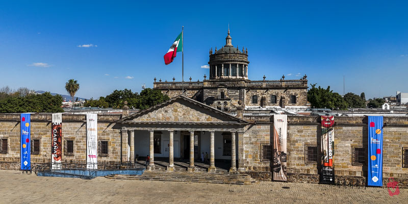 The Cabañas Museum, a historic building in Guadalajara, with a large Mexican flag flying above it against a clear blue sky.