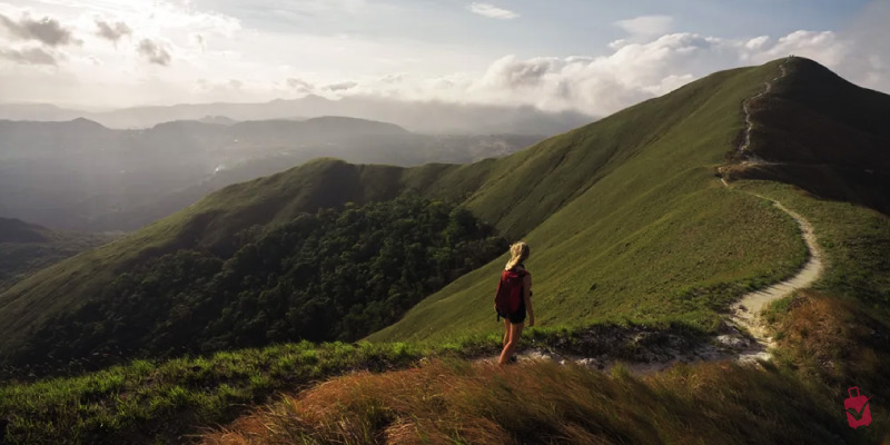 A person with a red backpack hiking on a grassy mountain ridge of Cerro de la Silla, with a winding path and distant mountains under a cloudy sky.