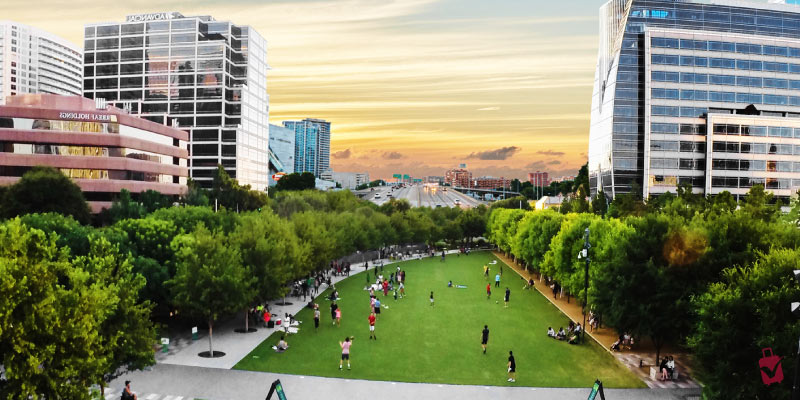People relax on the lush green grass of Klyde Warren City Park in Dallas which is nestled between modern skyscrapers and leafy trees at dusk.