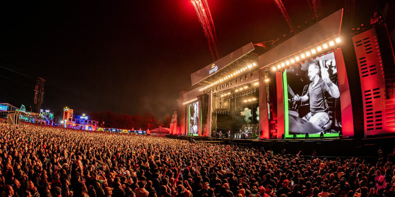 A massive outdoor music concert at night with a large crowd facing a brightly lit stage with pyrotechnics.