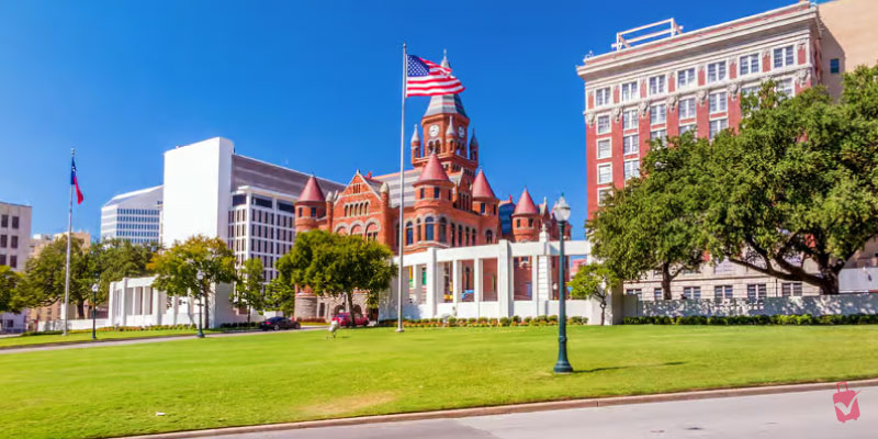 Lush green lawns and white pergolas at Dealey Plaza lead the eye toward the historic brick building housing The Sixth Floor Museum.