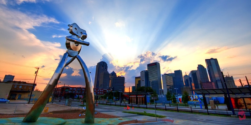 The tall silver Traveling Man sculpture stands in the Deep Ellum district of Dallas against a sunset sky featuring dramatic sun rays over the city.