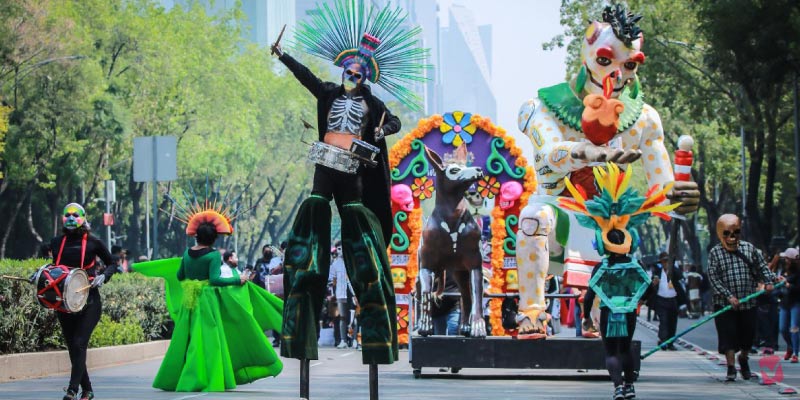 A vibrant Day of the Dead parade with people in skeleton costumes on stilts and large colorful floats.