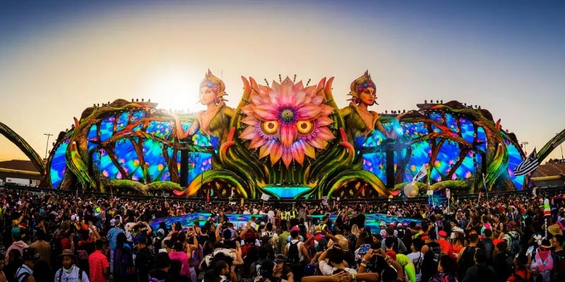 A large music festival stage elaborately decorated with colorful, nature-inspired designs and a huge crowd.
