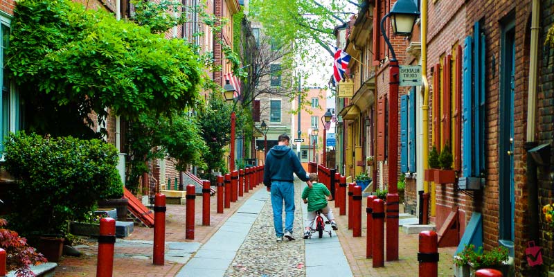 A man and child on a bicycle walk down the historic cobblestone path of Elfreth’s Alley lined with brick colonial houses.
