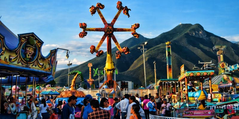 A lively amusement park scene at the Expo Feria Guadalupe, with various rides, including a spinning octopus-like ride and a carousel, set against mountains.