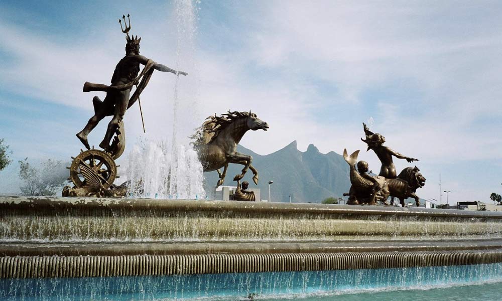 The Fuente de Neptuno fountain, a famous historical landmark in Monterrey, Mexico, with bronze statues of Neptune and horses spouting water, against a mountain backdrop.
