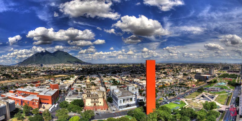 A panoramic view of Monterrey, Mexico, with the tall orange Faro de Comercio skyscraper prominently in the foreground, and mountains under a blue sky.