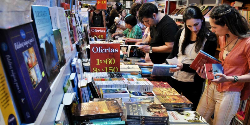 A busy book fair scene with many people browsing books on shelves and tables.