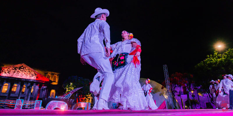 Traditional Mexican dancers in white attire perform on an outdoor stage at night during the Festival Cultural de Mayo in Guadalajara.