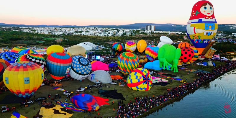 A panoramic view of a hot air balloon festival with dozens of colorful balloons of various shapes and sizes near a body of water and a city.
