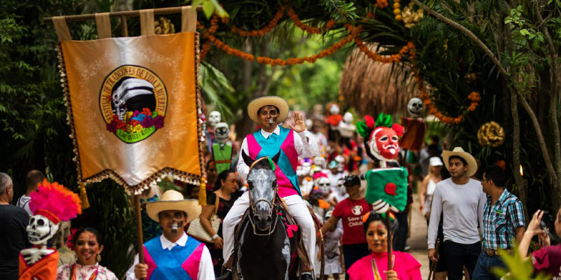 A festive parade with a person on horseback waving to the crowd, surrounded by people in traditional Mexican attire and a large banner.