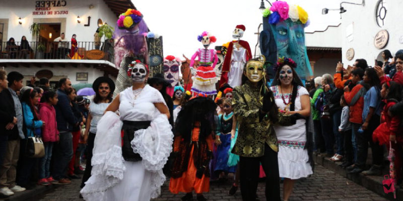 A Day of the Dead parade with people in elaborate skull makeup and traditional attire walking down a street with spectators.
