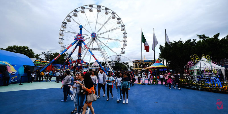 Crowds walk through a vibrant fairground featuring a large white Ferris wheel and colorful rides during the Fiestas de Octubre in Guadalajara.