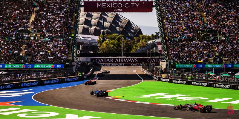Formula 1 race cars speeding around a track in Mexico City, surrounded by a grandstand filled with cheering spectators.
