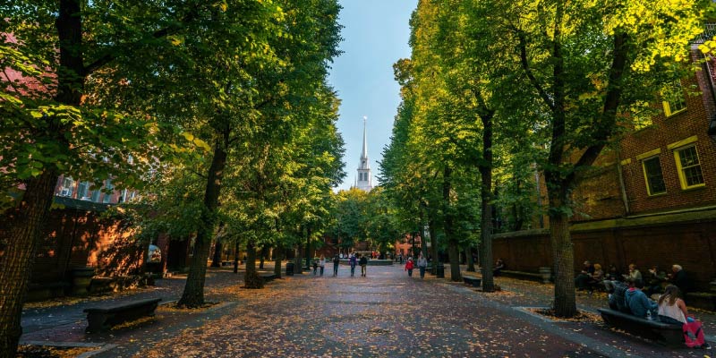 A brick walkway lined with trees leads toward the Old North Church spire, representing a key stop along the iconic Freedom Trail walking tour in Boston.