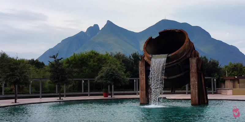 A large, rusty metal sculpture shaped like a pouring pot, with water cascading into a pool at Fundidora Park, Monterrey, Mexico, with mountains in the background.