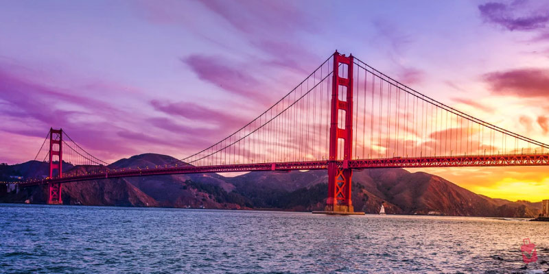 The iconic Golden Gate Bridge stands majestically over the water against a stunning purple and orange sunset sky with rolling hills in the distance.