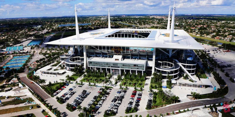 An aerial view of Hard Rock Stadium in Miami, surrounded by parking lots and some greenery.
