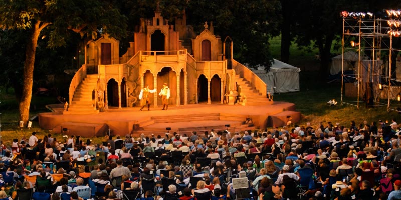 A large crowd watches two actors perform on a lit outdoor stage with a castle backdrop during the Heart of America Shakespeare Festival.